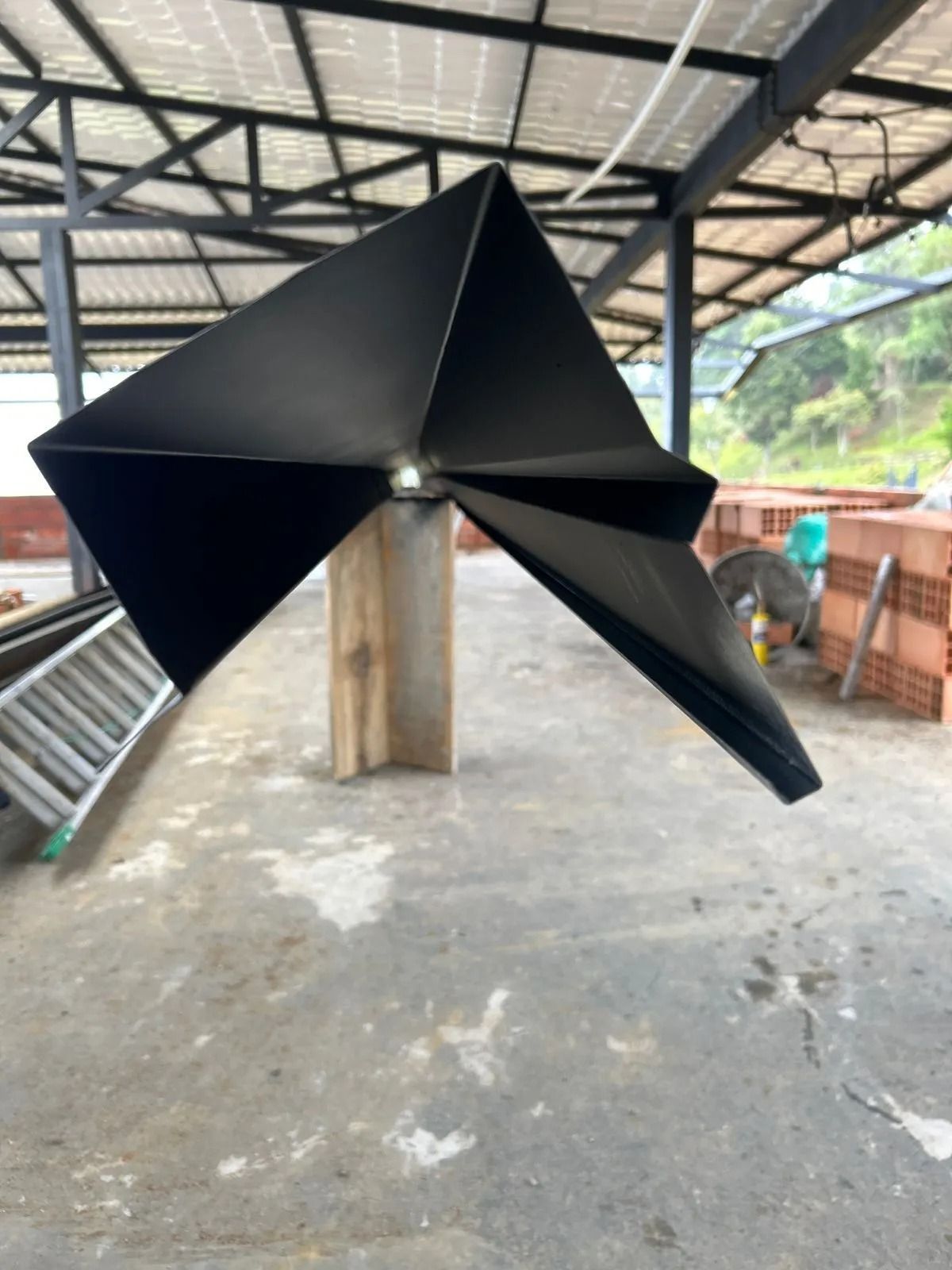 Escultura geométrica de metal negro sobre una base de hormigón en un taller al aire libre.