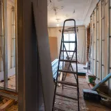 Narrow hallway under renovation with ladder, exposed walls, and construction materials scattered on the floor