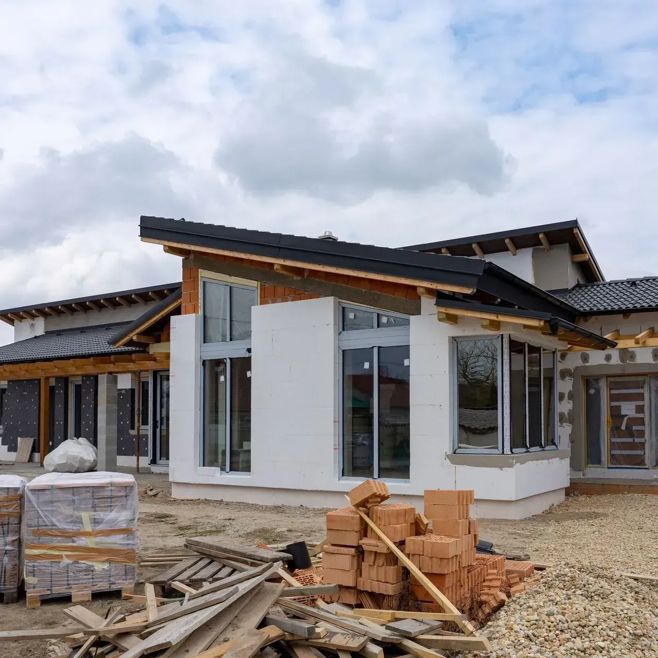 Partially built modern house with white walls, dark roof, and construction materials in the foreground