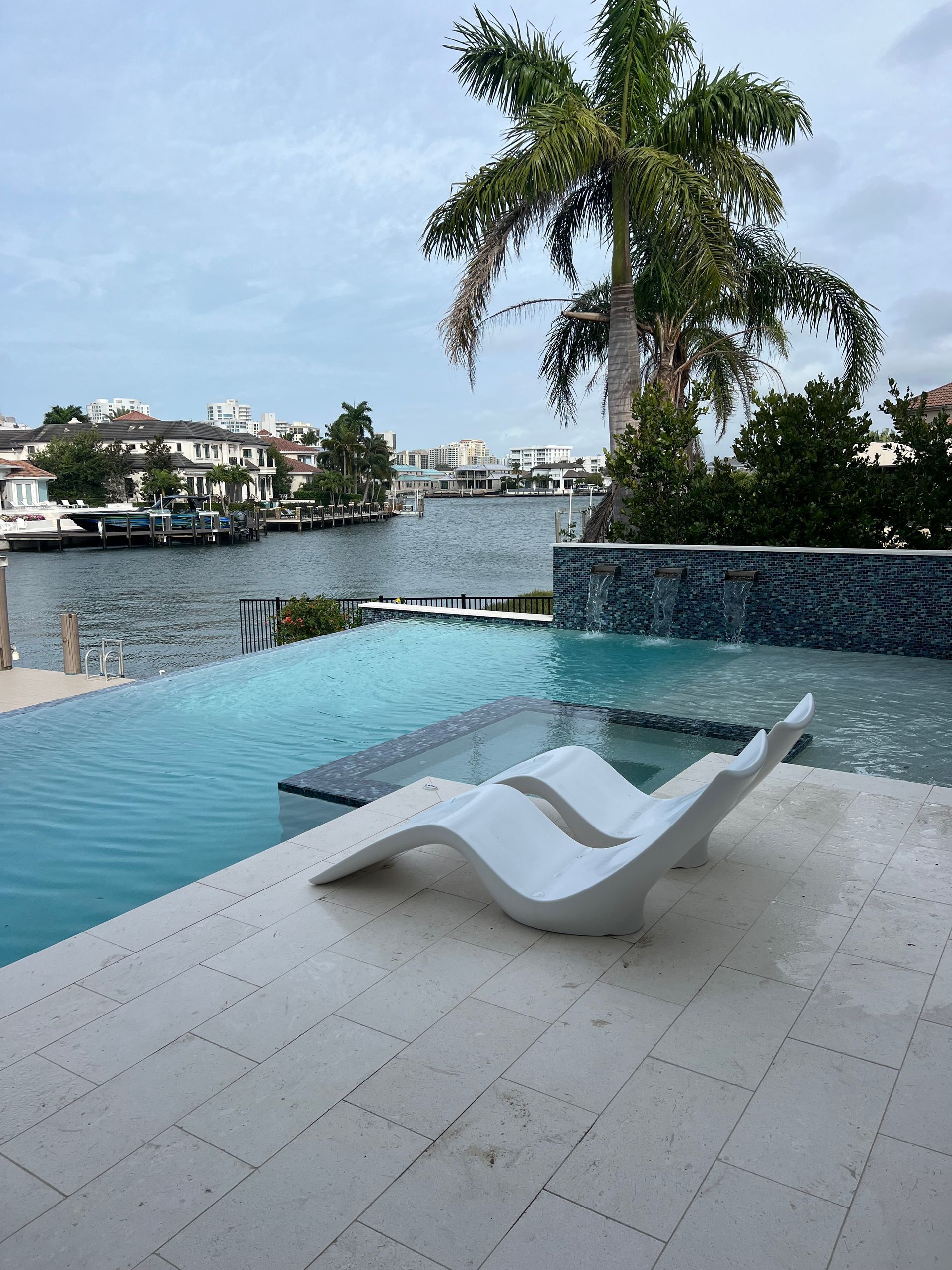 Infinity pool with white lounge chair overlooking a calm waterfront and palm trees at sunset