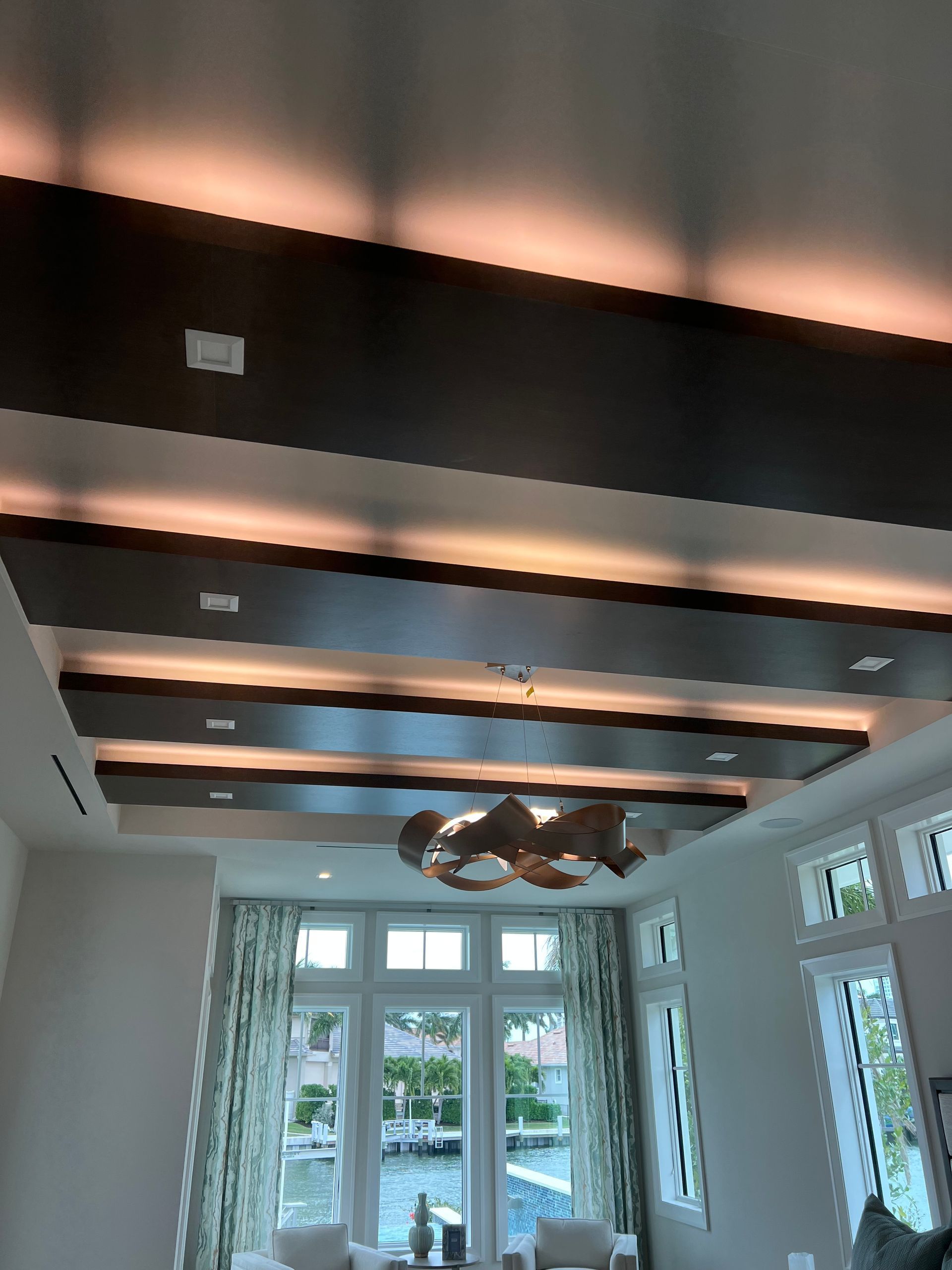 Modern bright room with a dark beam ceiling, chandelier, and large windows opening to a patio and greenery