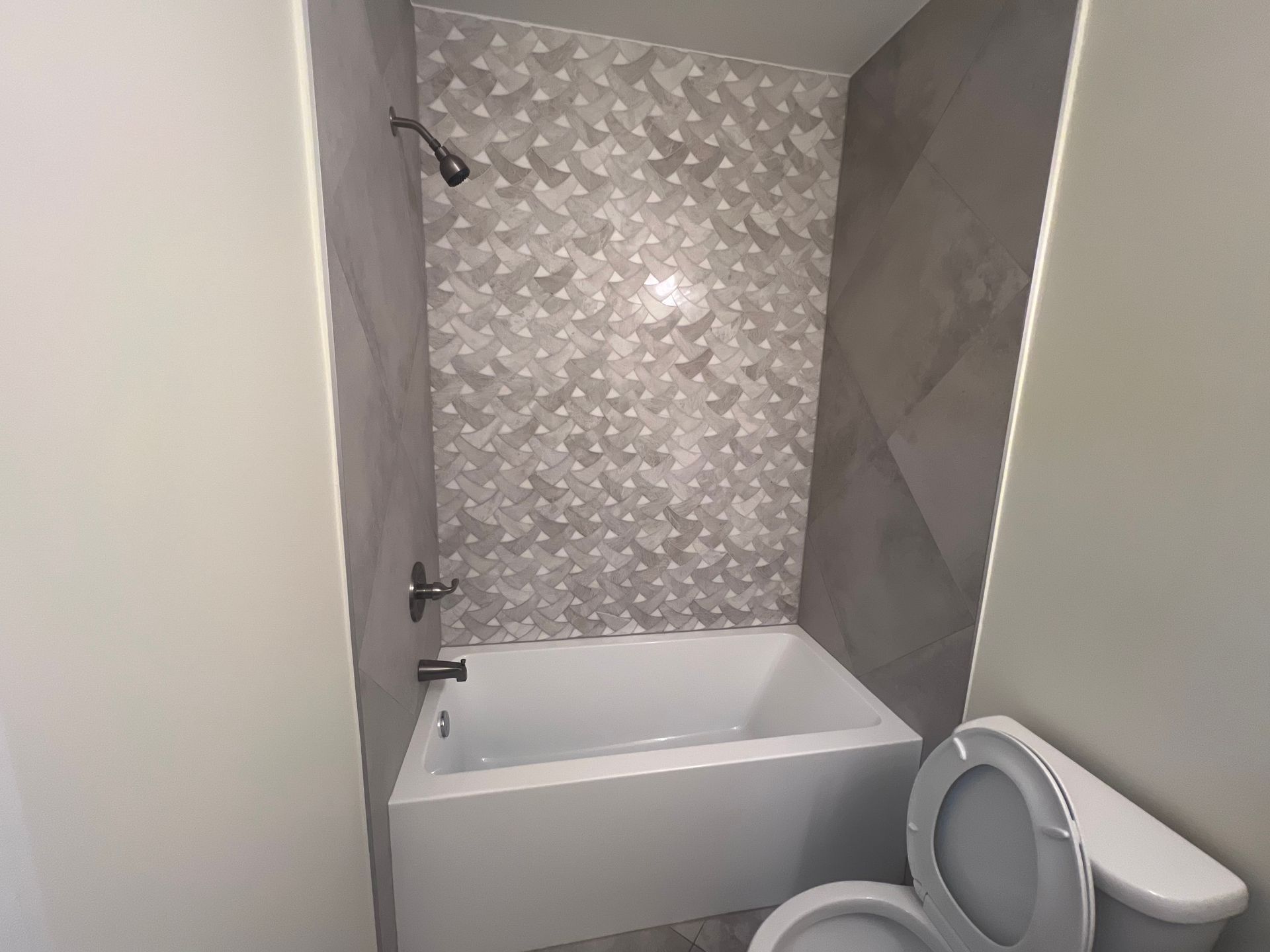 Small bathroom with a white bathtub, gray tile walls, and a toilet in the foreground.