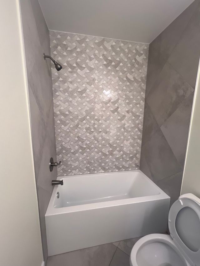 Small bathroom with a white tub, gray tiled walls, and a partial toilet in the foreground