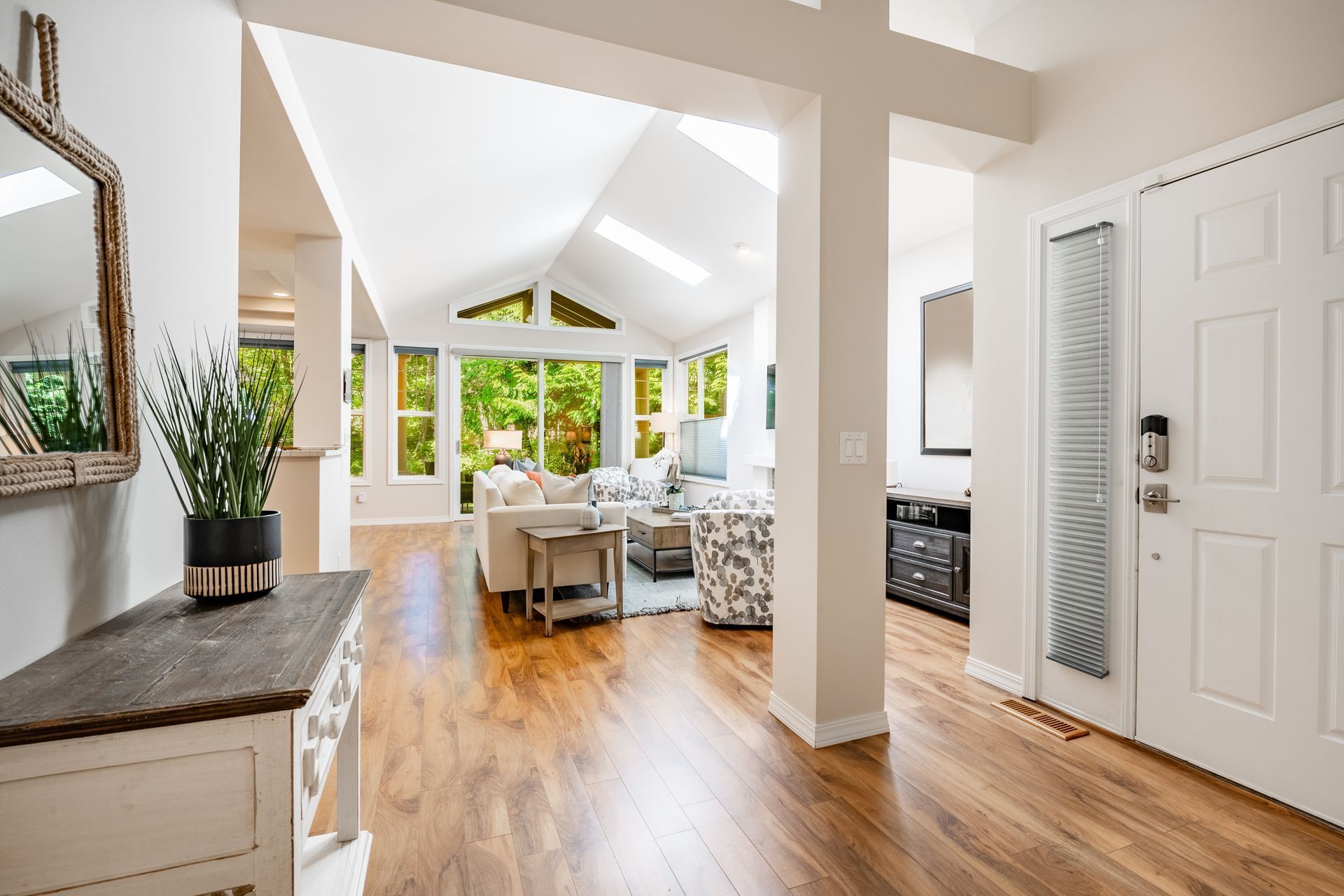 Bright open living room with vaulted ceiling, wood floors, white furniture, and large windows overlooking greenery