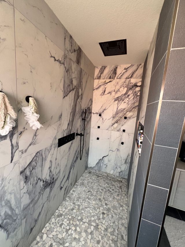 Narrow marble-tiled bathroom or shower corridor with pebble floor and black fixtures, unfinished with ceiling vent