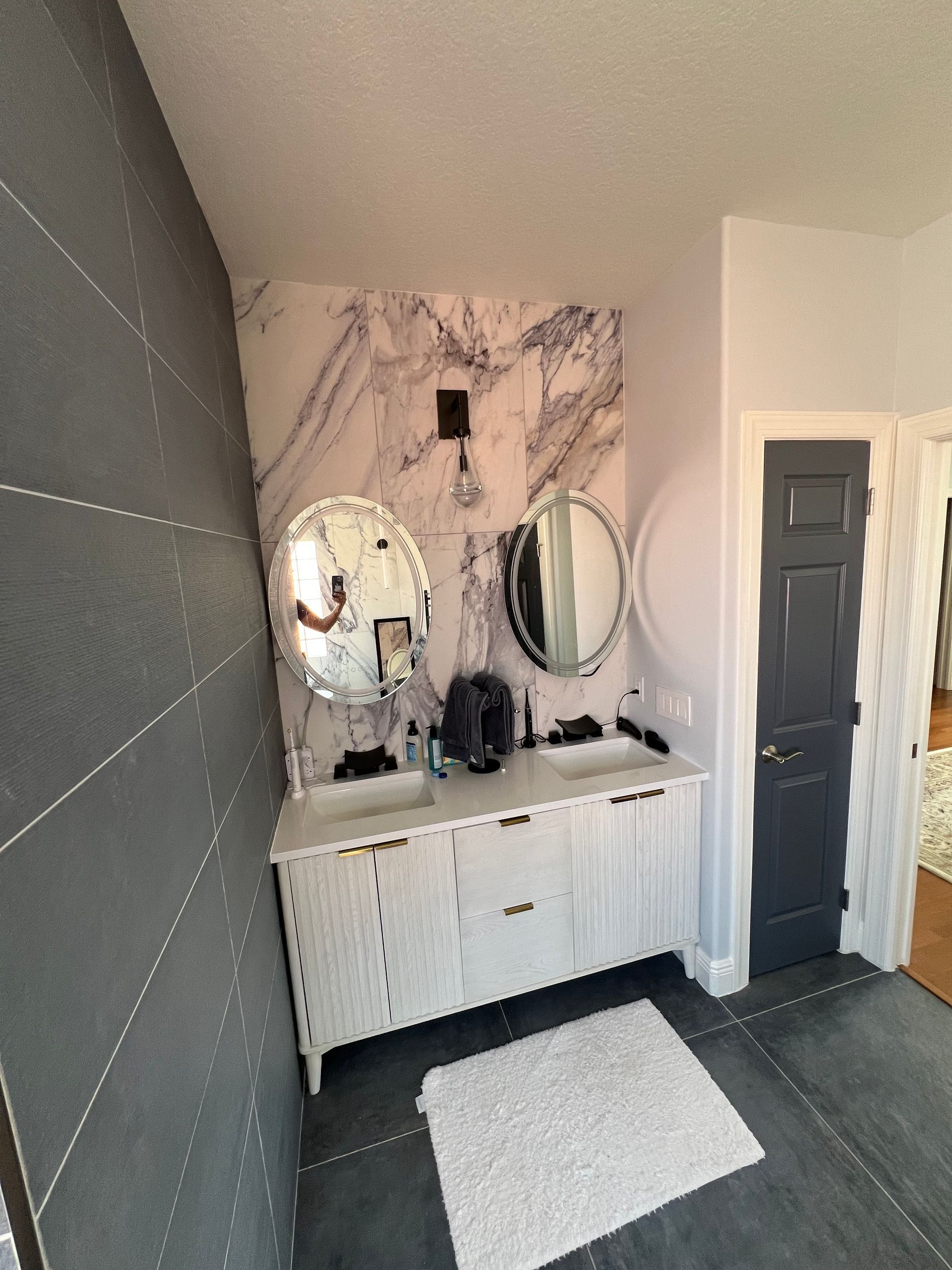 Modern bathroom vanity with dual mirrors, marble backsplash, dark tiled floor, and white bath mat
