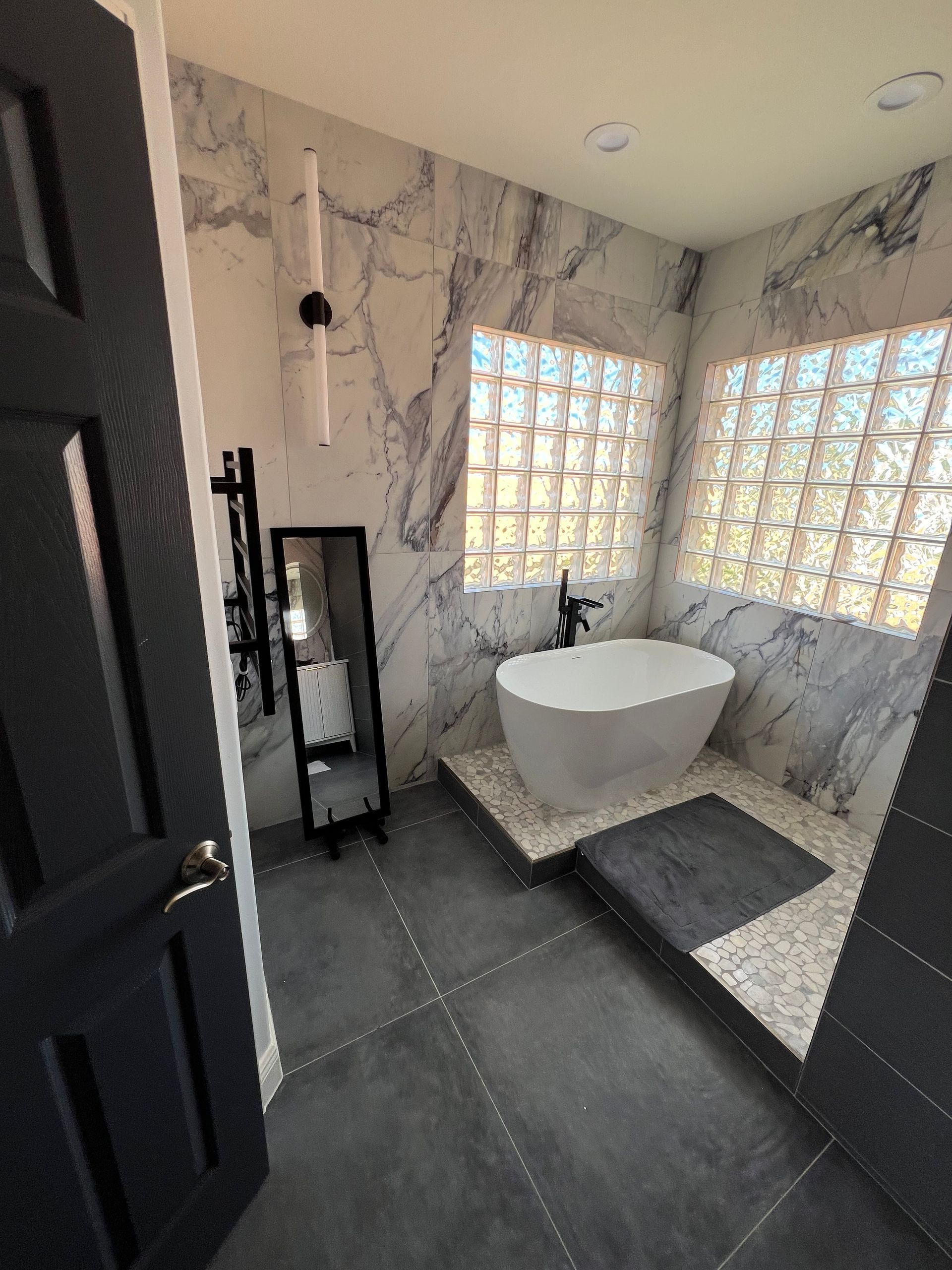Modern bathroom with freestanding tub, marble-look walls, dark floor, and glass block windows