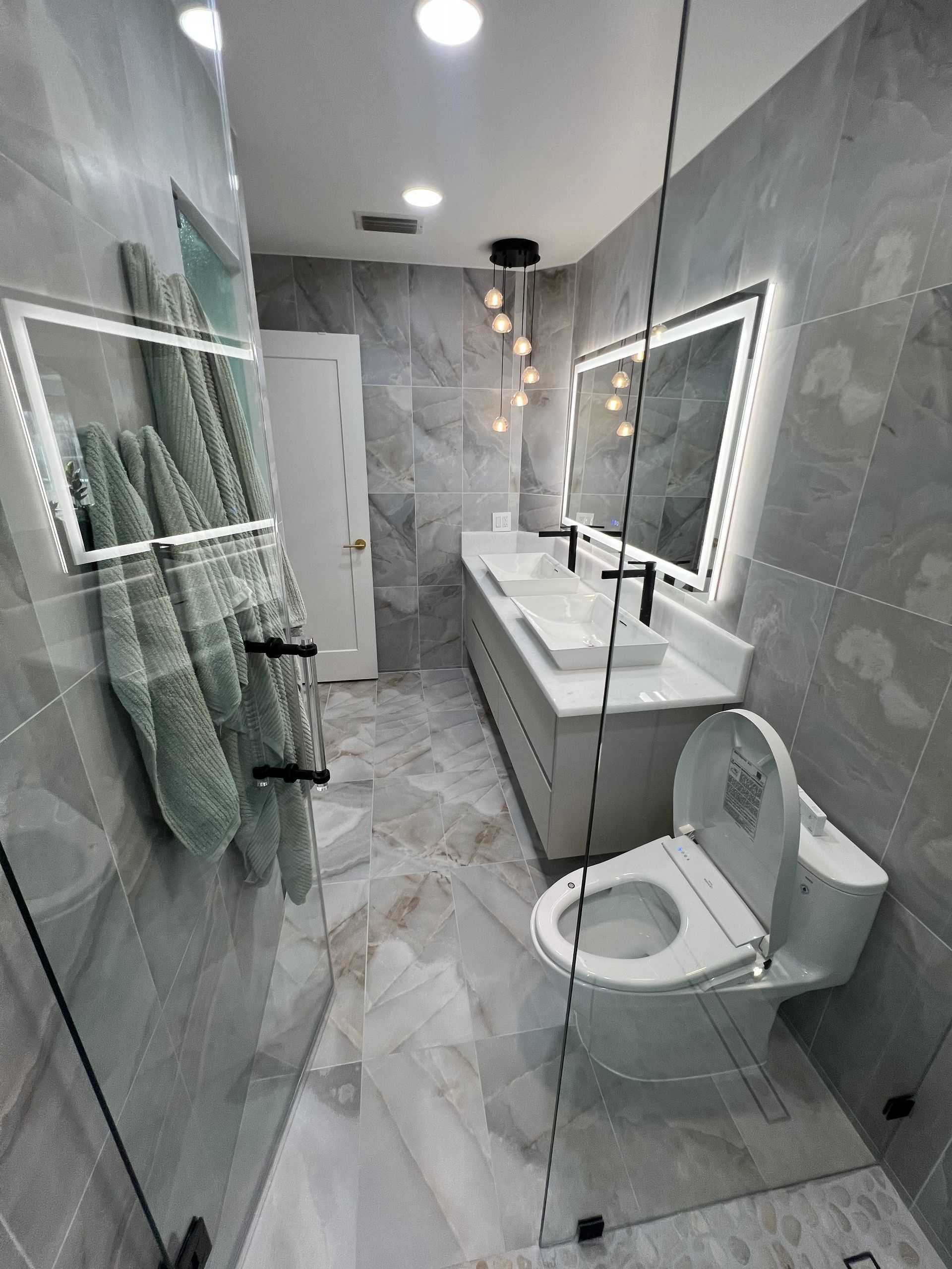 Modern bathroom with glass shower, white sink, wall mirror, and toilet on marble tile floor