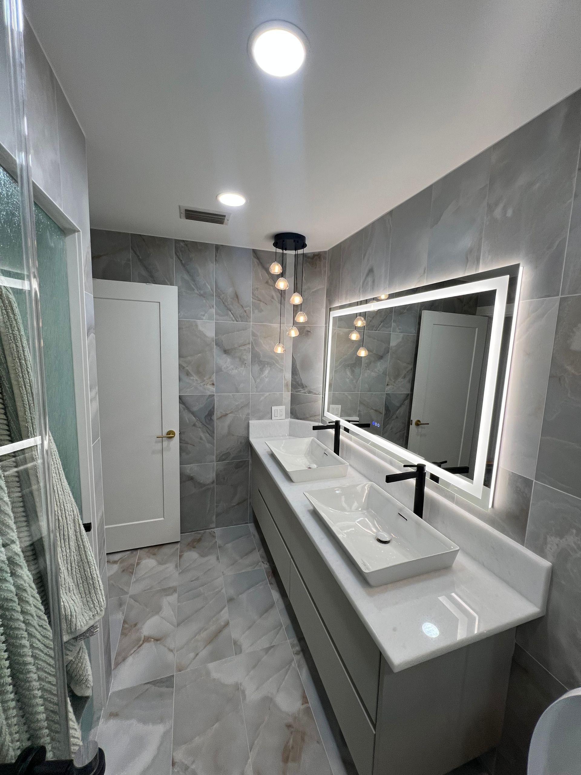 Modern bathroom with double vanity, large lit mirror, gray tile walls, and glass shower enclosure