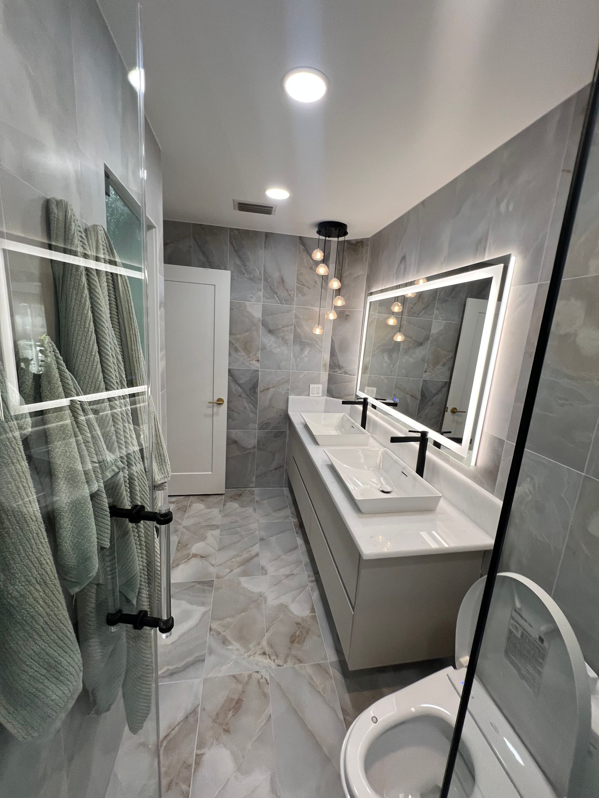 Modern bathroom with double vanity, large mirror, glass shower, and gray tile flooring.