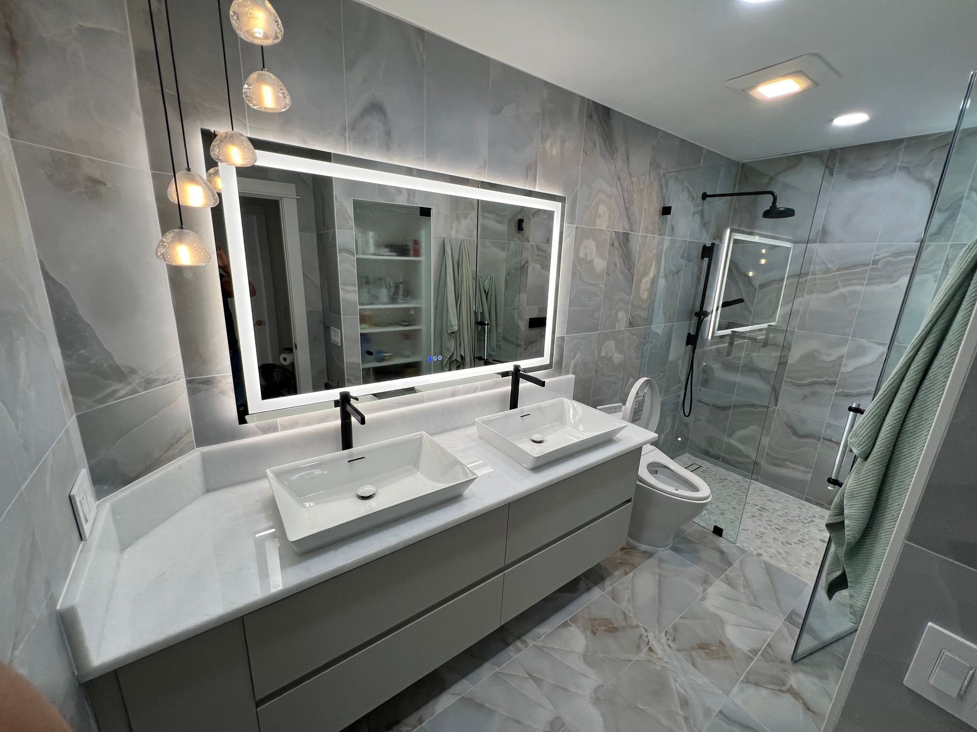 Modern gray bathroom with double vanity, backlit mirror, glass shower, and towel hanging on the wall