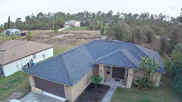 Aerial view of a suburban house with a blue roof and a front porch, surrounded by trees and neighboring homes.