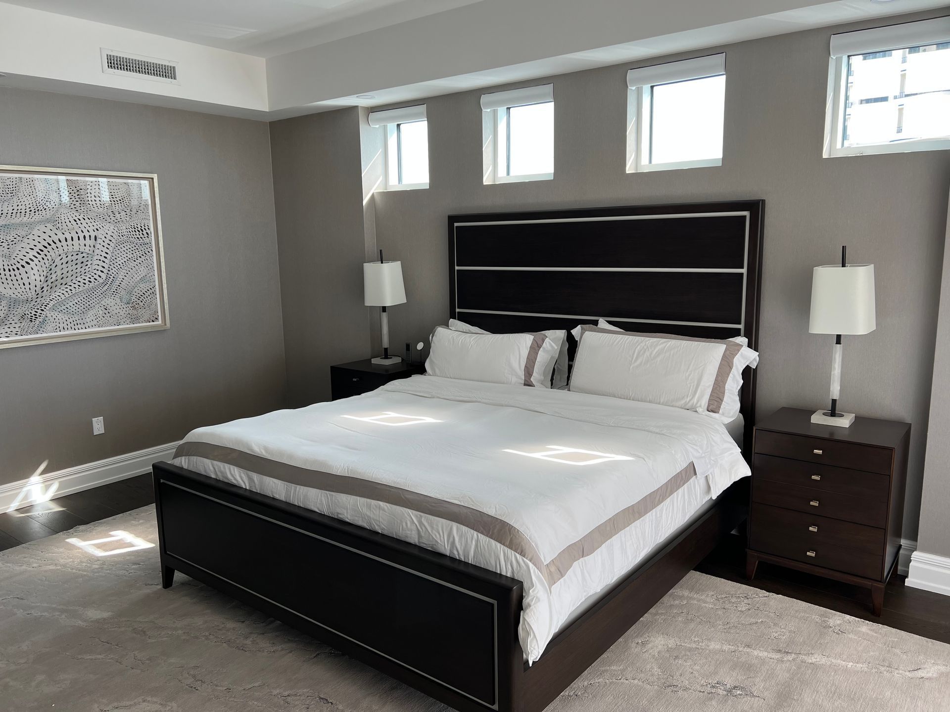 Modern bedroom with a large bed, dark wood furniture, and high windows letting in natural light.