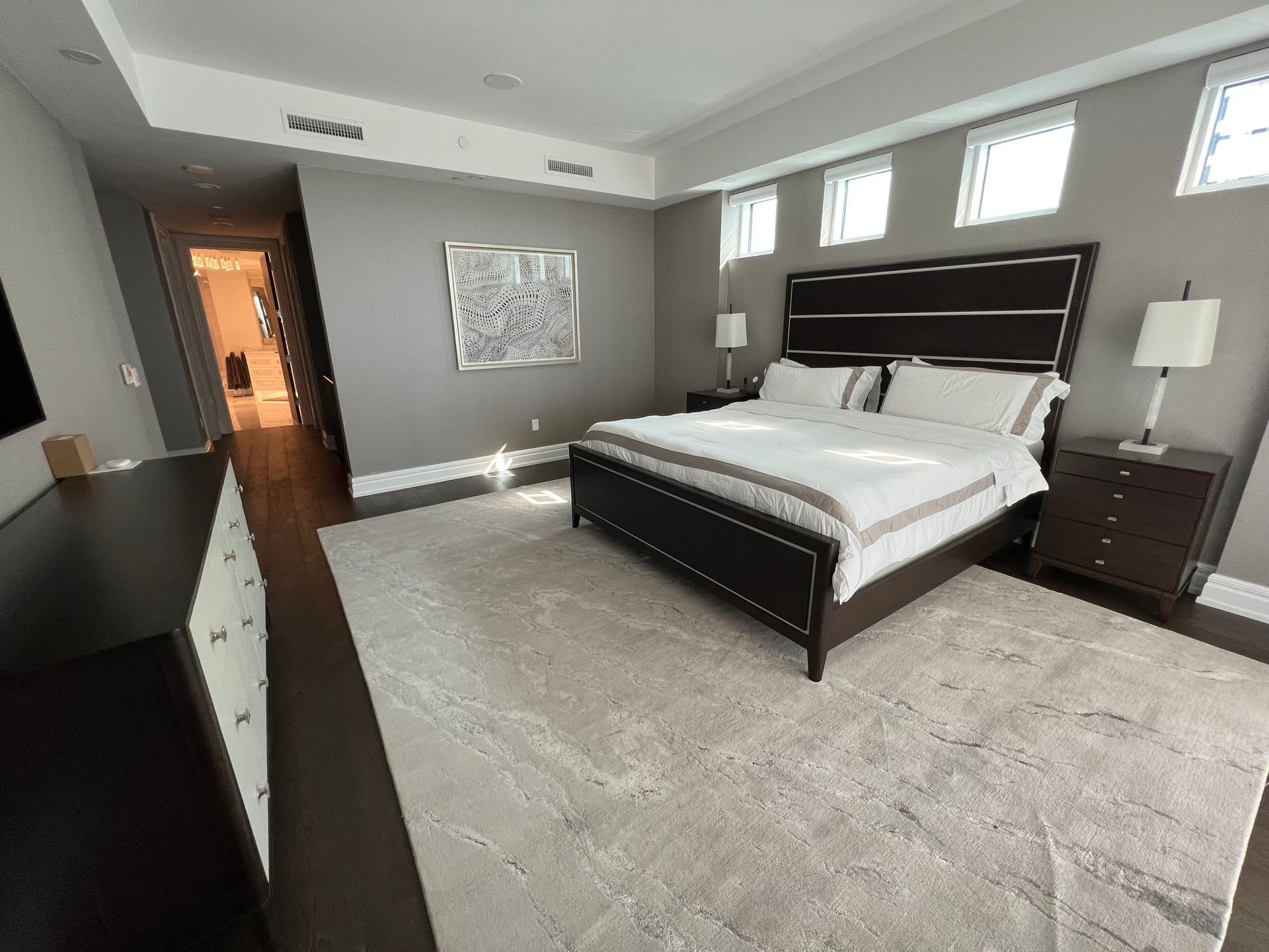 Modern bedroom with a dark wood bed, gray walls, carpet, and three small windows above the headboard