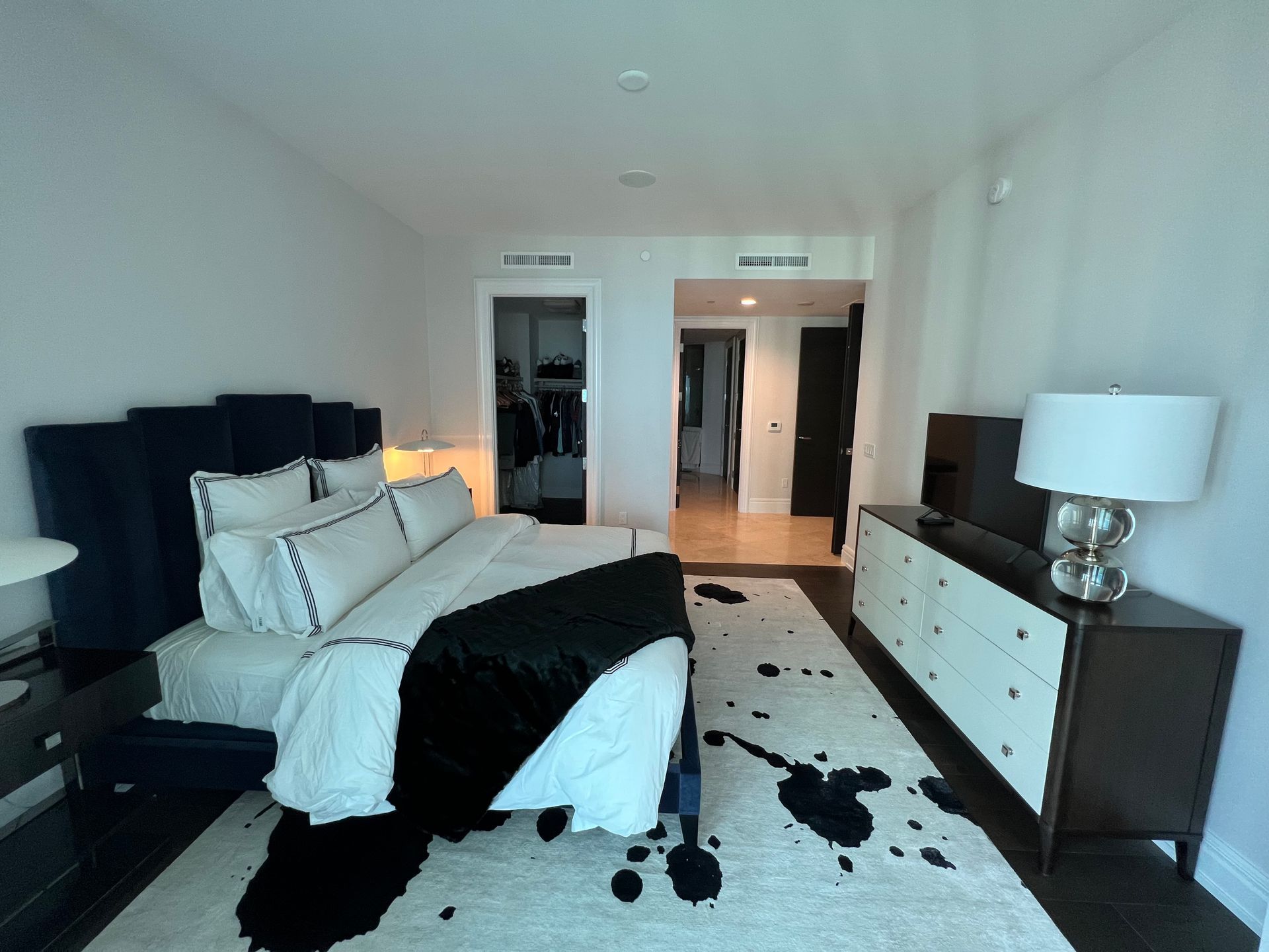 Bright modern bedroom with white bedding, black accents, patterned rug, and a dresser with lamp