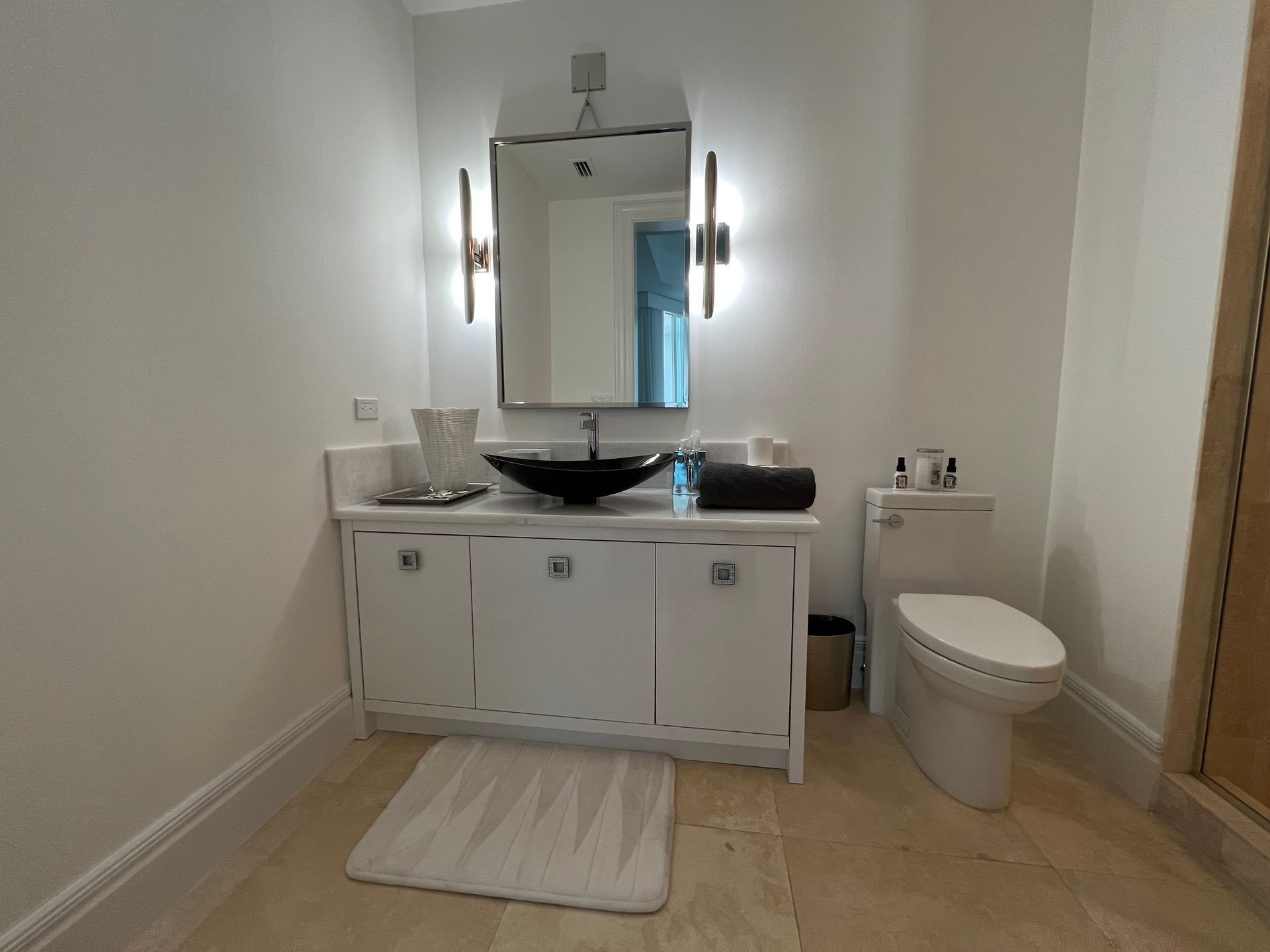 Small bathroom vanity with mirror, black sink, white cabinets, toilet, and light rug.