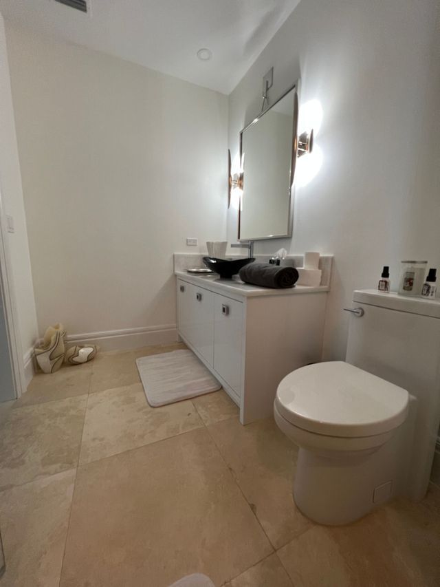 Small white bathroom with double sink vanity, mirror, toilet, rug, and beige tiled floor