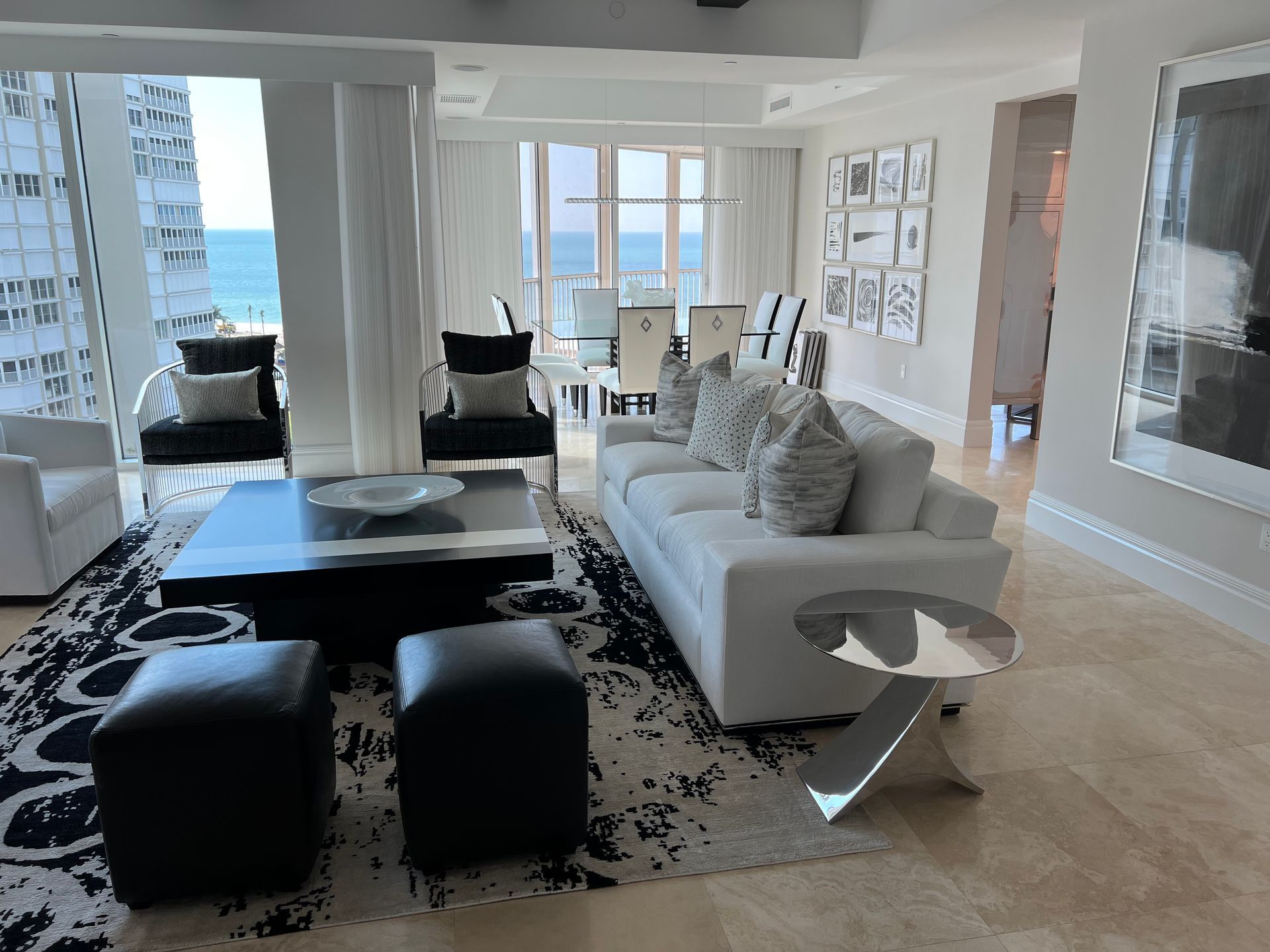Modern living room with white sofa, black ottomans, patterned rug, and ocean view through large windows