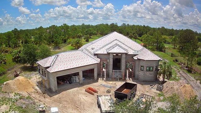 Partially built house with a light roof and open garage, surrounded by cleared land and trees under a blue sky