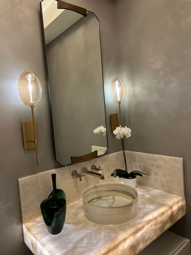 Small powder room vanity with brass wall sconces, round mirror, stone sink, and dark green vase.