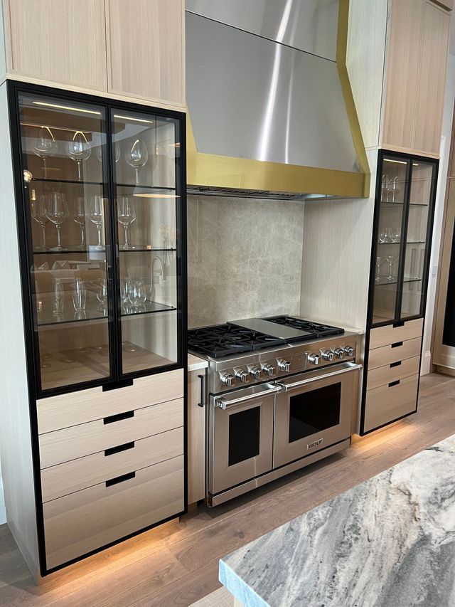 Modern kitchen with stainless steel range, gold hood, and black glass-front cabinets; marble island edge in foreground
