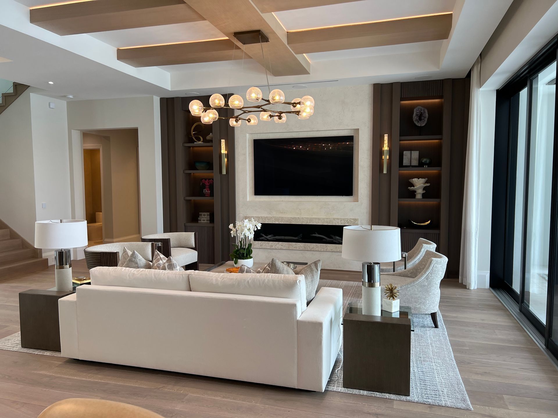 Modern living room with white sofas, wall-mounted TV, chandelier, and floor-to-ceiling windows