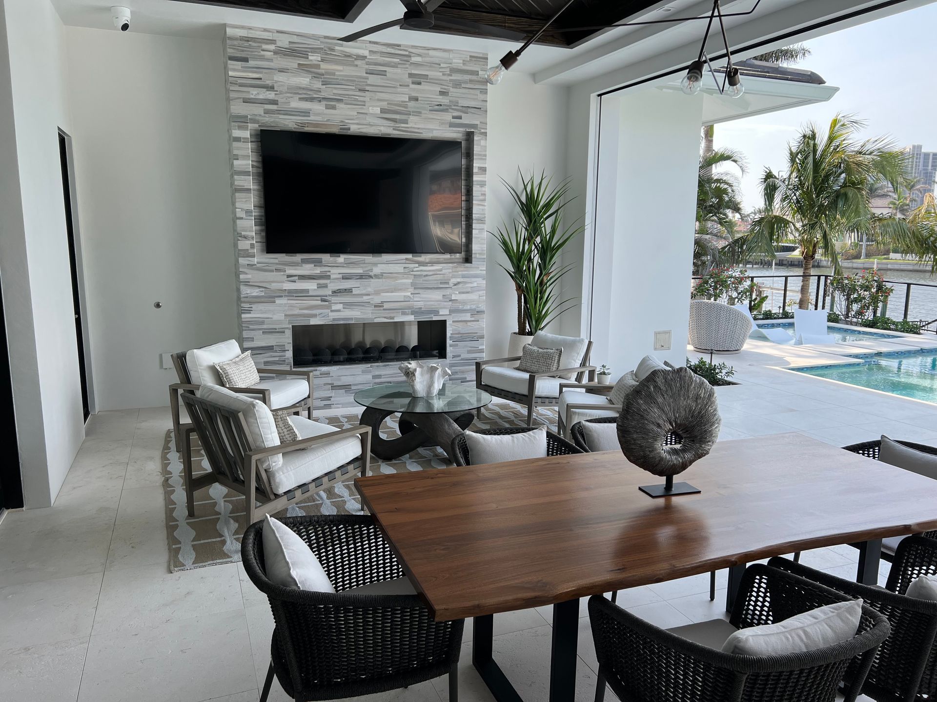 Modern patio lounge with dining table, wicker chairs, wall-mounted TV, fireplace, and pool view