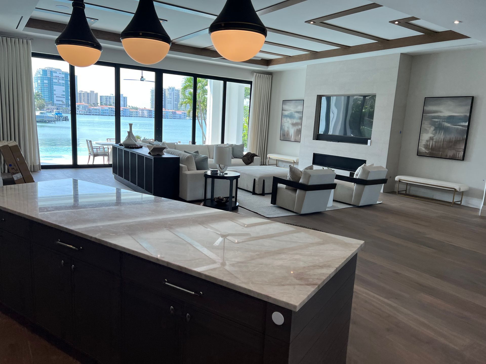 Modern living room with white sofas, dark wood floors, marble kitchen island, and large ocean-view windows