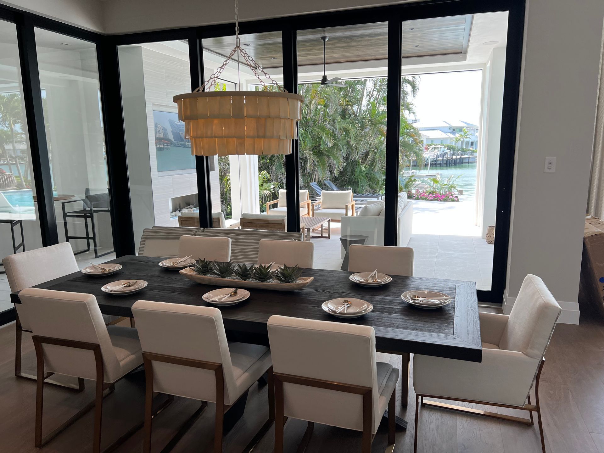 Modern dining room with a long dark table, beige chairs, and large glass doors opening to a patio and water view