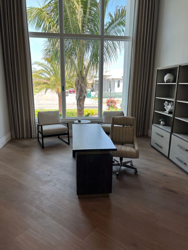 Modern home office with large windows, desk, chair, armchair, and shelving facing palm trees outside