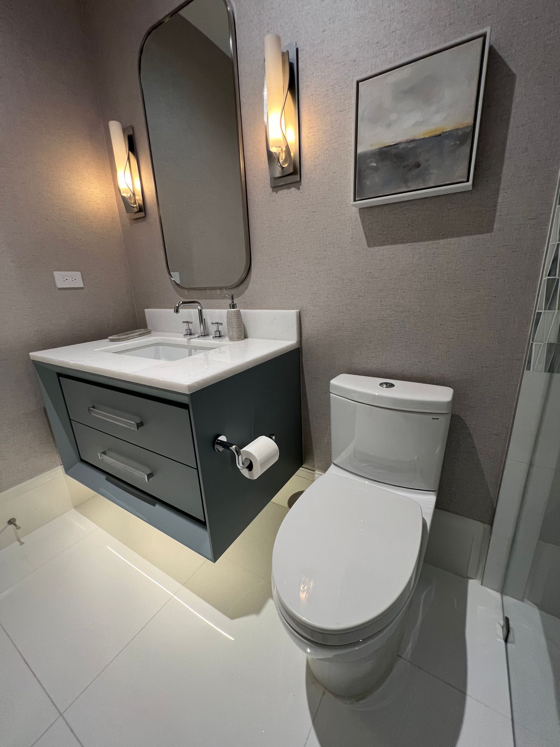 Small bathroom with gray vanity, mirror, wall lights, and toilet on a white tile floor
