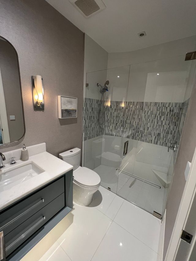 Modern bathroom with white tile, gray vanity, toilet, and glass-enclosed shower with mosaic accent wall