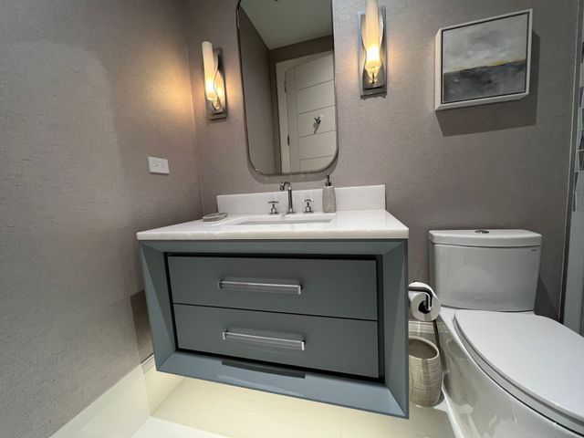 Small modern bathroom with gray vanity, white sink, mirror, wall sconces, toilet, and framed art.