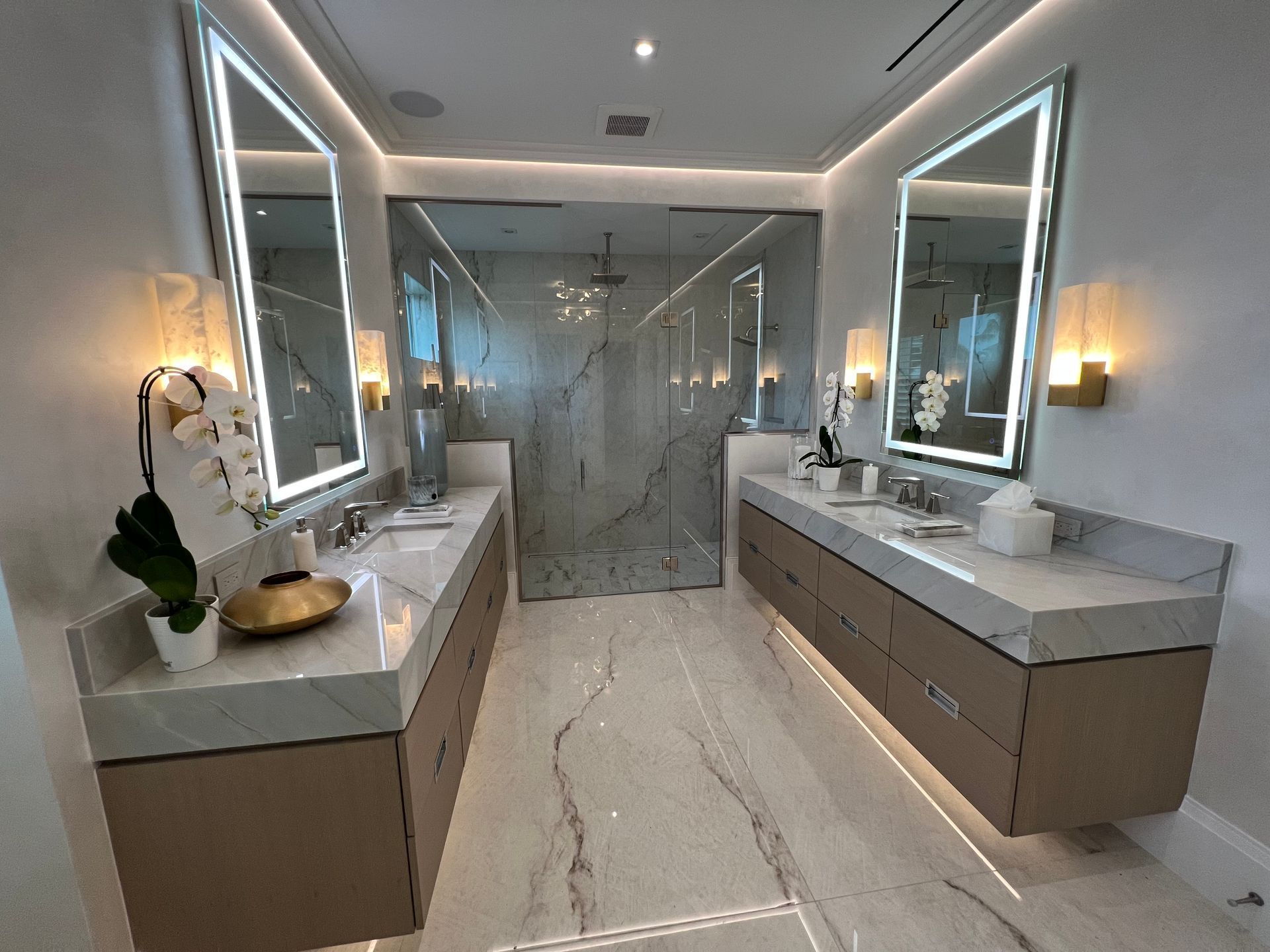 Modern double vanity bathroom with lit mirrors, gray marble surfaces, and glass shower enclosure