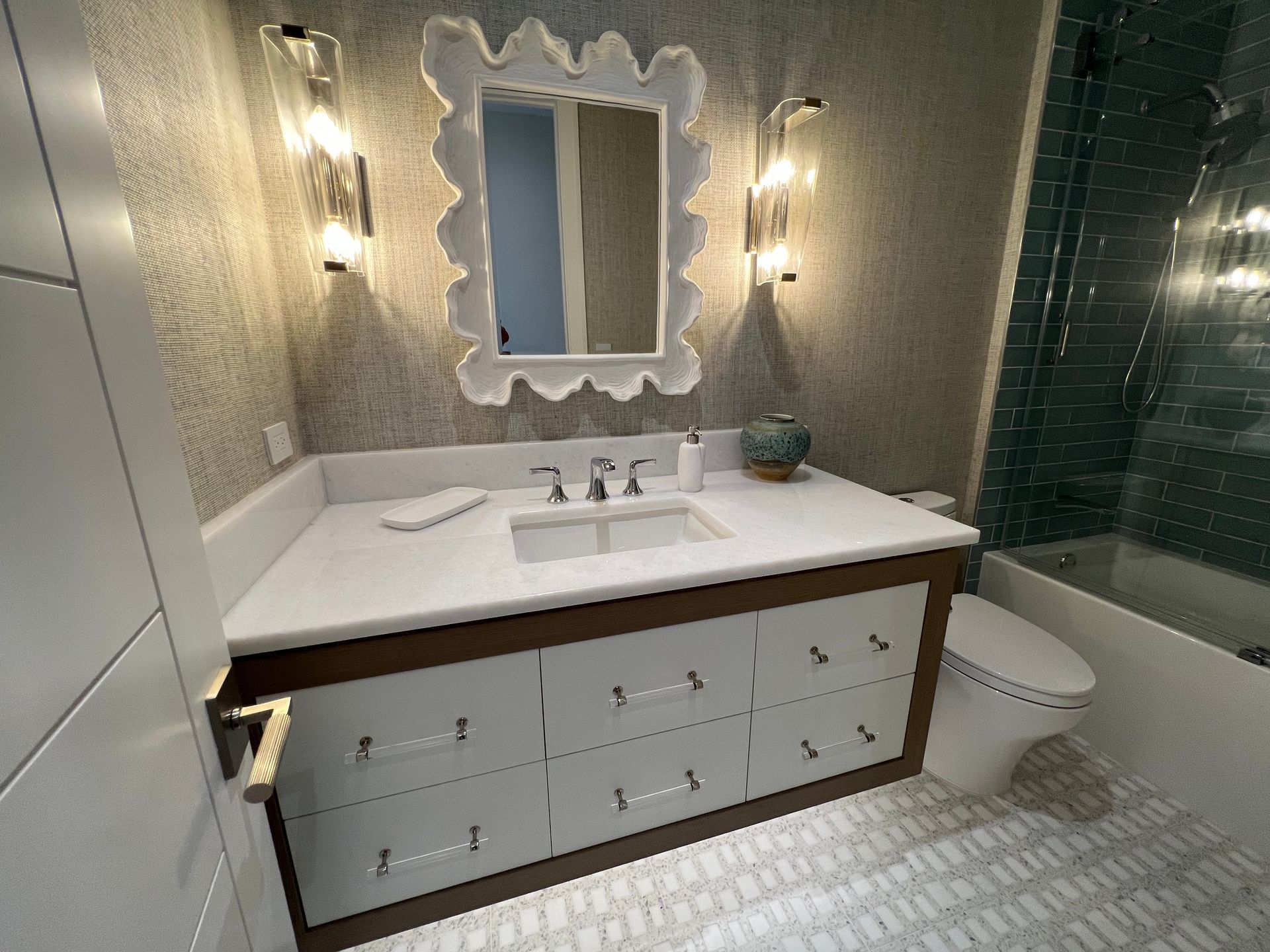 Modern bathroom vanity with white countertop, ornate mirror, wall sconces, and glass shower enclosure.