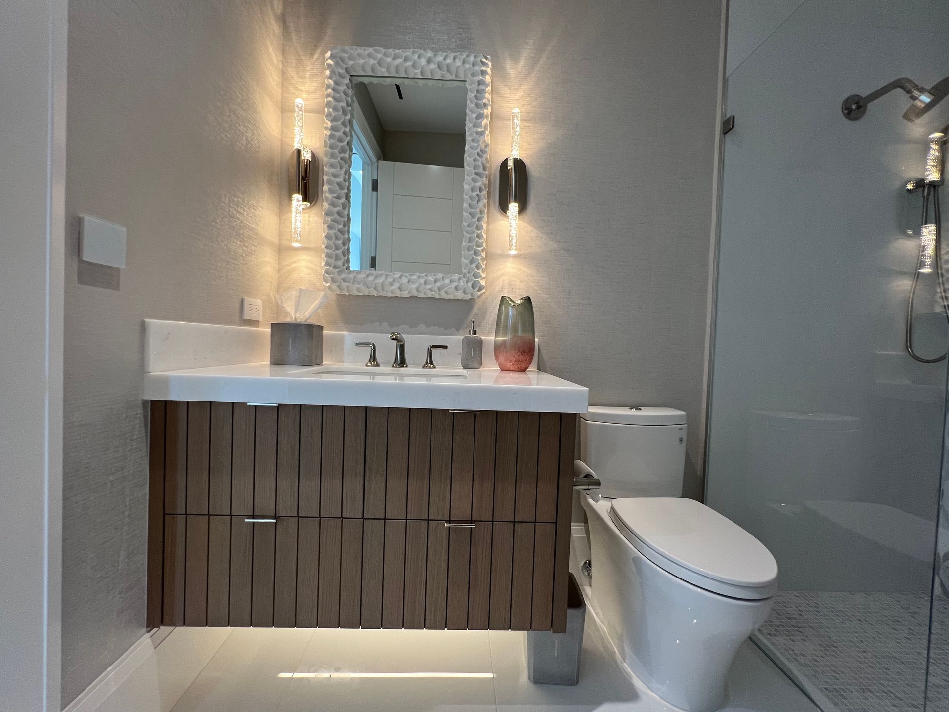 Modern bathroom with floating wood vanity, mirror, wall sconce, toilet, and glass shower enclosure