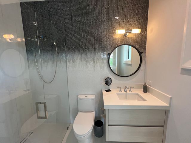 Modern bathroom with glass shower, toilet, vanity sink, round mirror, and dark textured accent wall