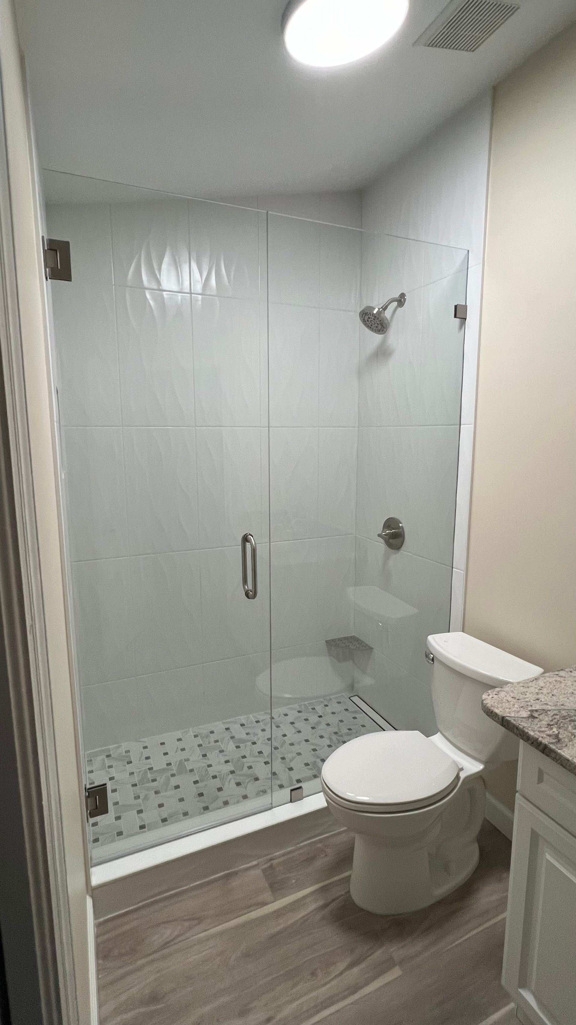 Modern bathroom with glass shower, white tile, and toilet beside a granite vanity.