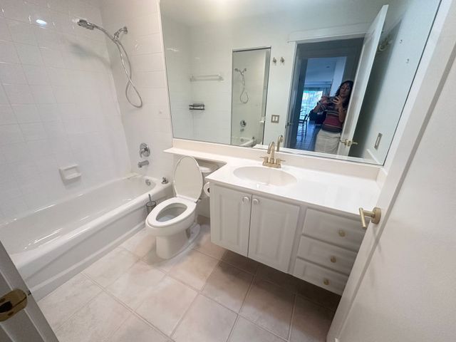 Bright bathroom with white tub, toilet, vanity, and large mirror; person visible in mirror doorway.