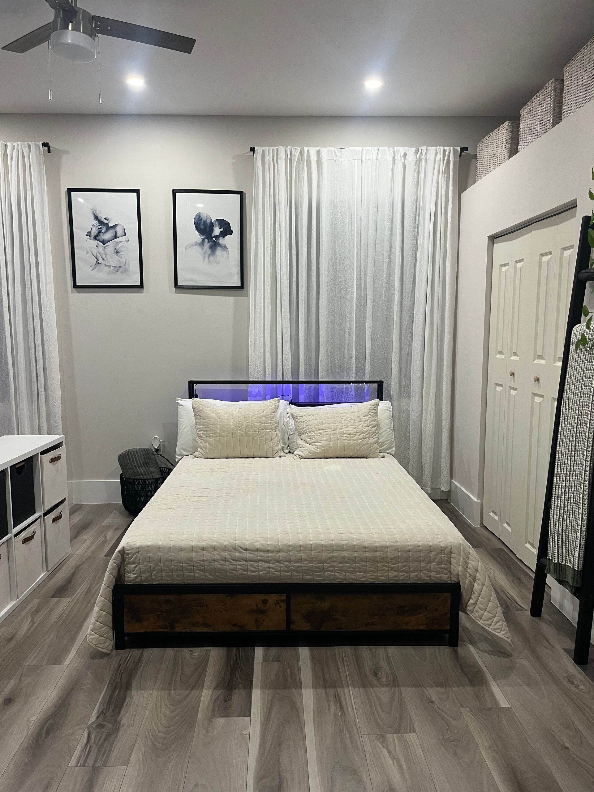 Bedroom with a neatly made bed, gray walls, white curtains, and framed artwork above the headboard