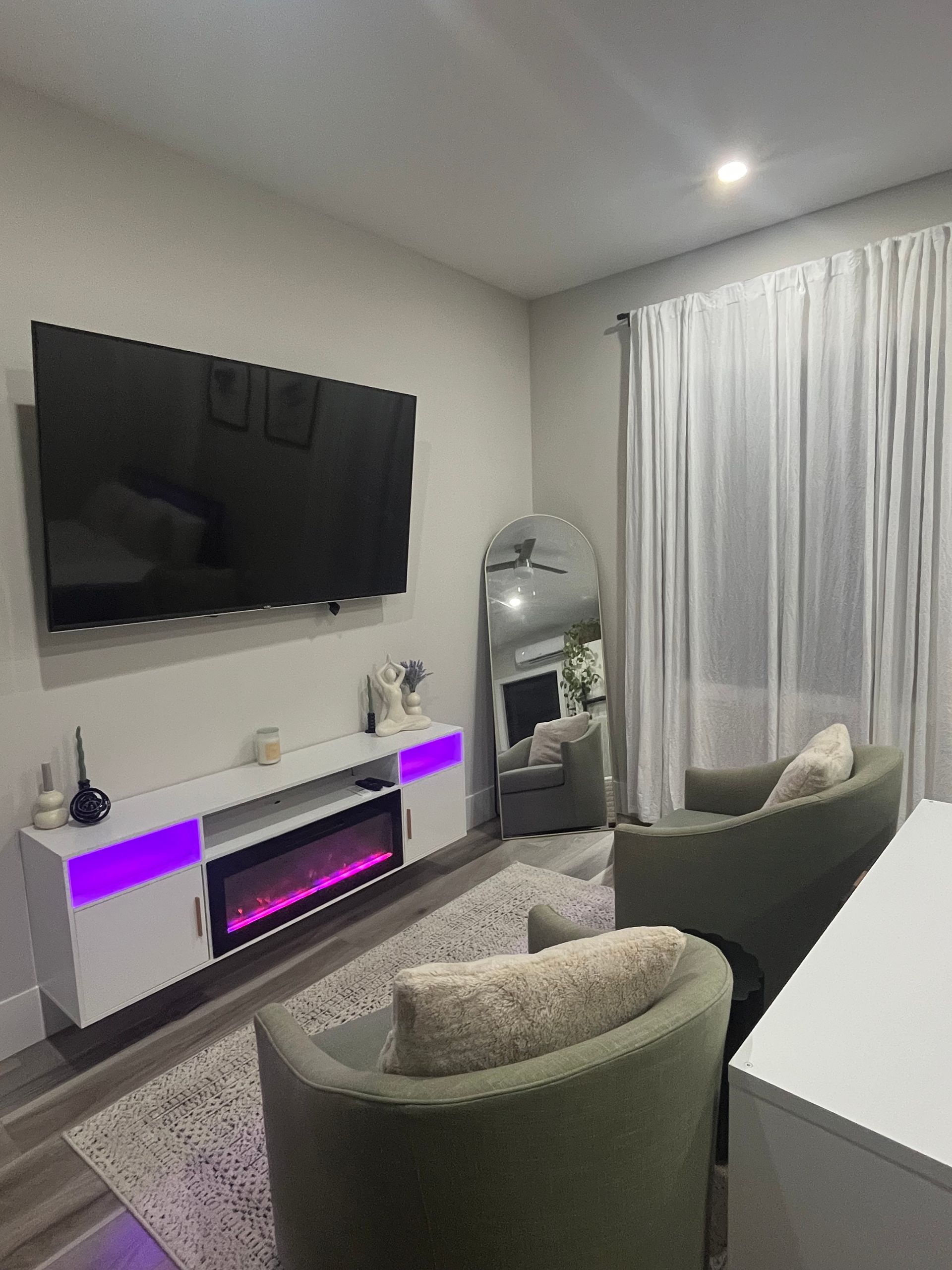 Modern living room with wall-mounted TV, purple-lit media console, armchairs, mirror, and curtains