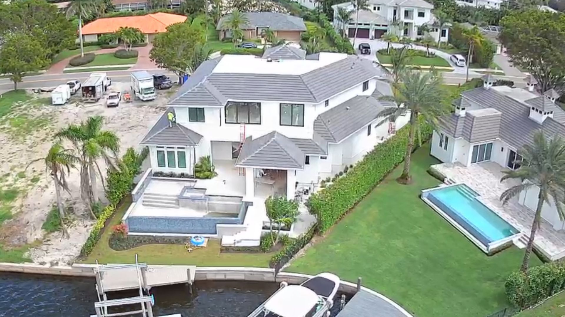 Aerial view of a large white waterfront house with a pool and dock beside neighboring homes.
