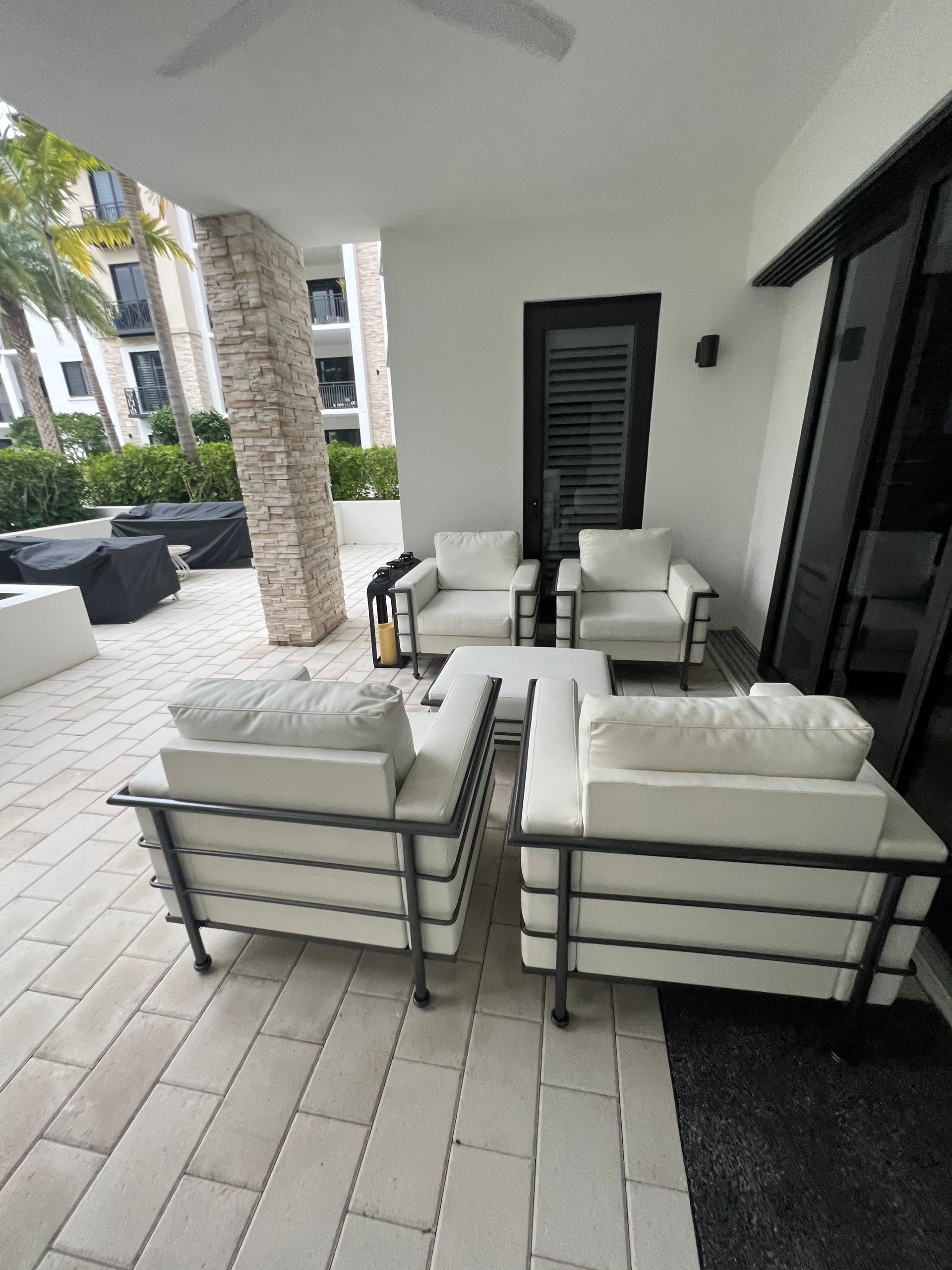 Covered patio seating area with white cushioned chairs and sofas beside glass doors and a poolside lounge area.