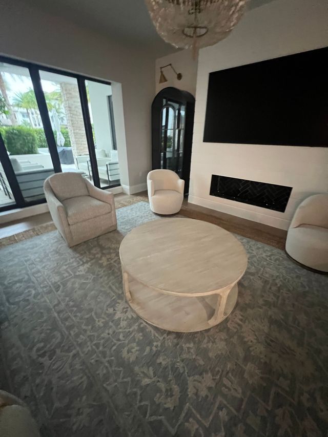 Modern living room with beige chairs, round coffee table, black fireplace, large wall-mounted TV, and sliding glass doors.