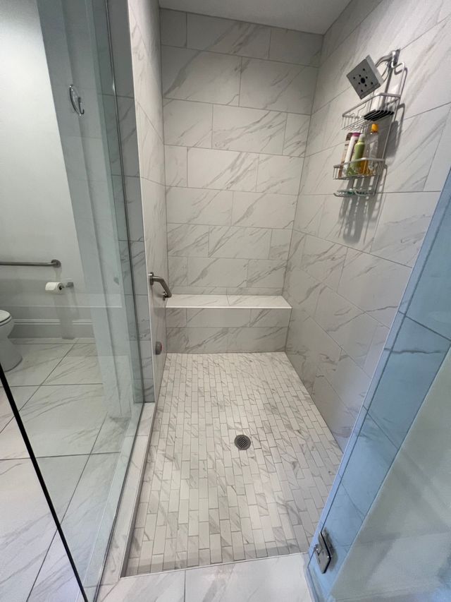 Modern white-tiled shower with glass door, bench, and wall-mounted caddy.