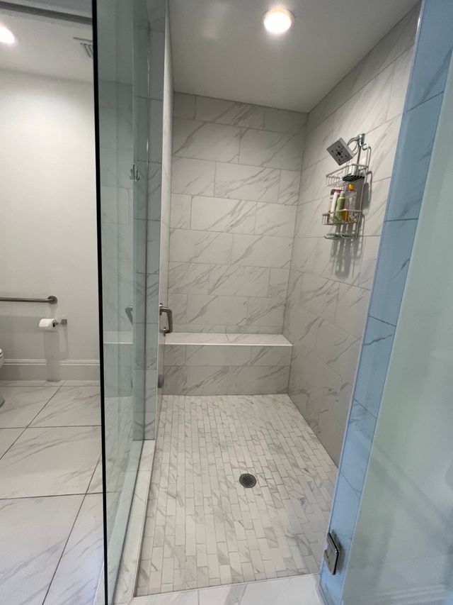 Modern tiled shower stall with glass door, built-in bench, and showerhead caddy