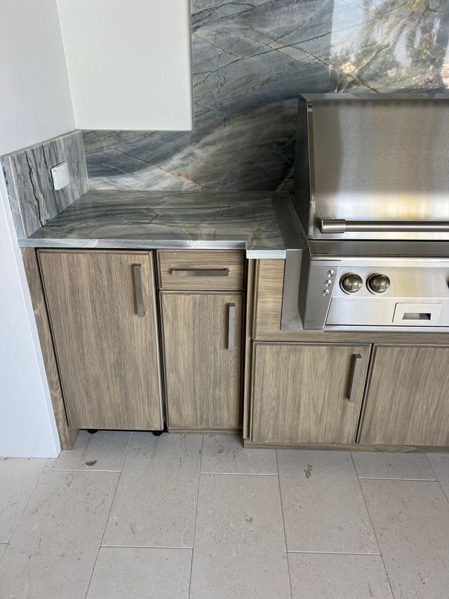 Outdoor kitchen cabinet and countertop with gray stone backsplash and built-in grill on tiled floor