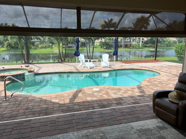 Covered backyard pool with brick patio, white chairs, blue umbrella, and a lake view beyond the screen enclosure.