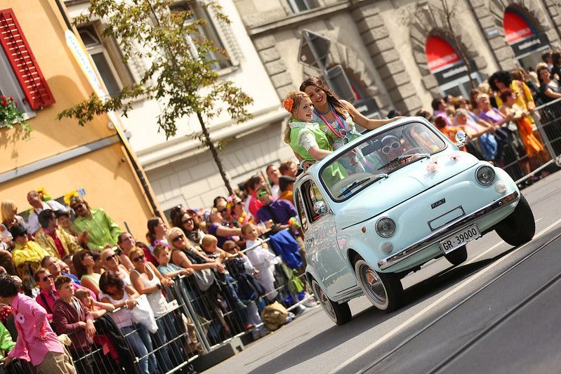Vintage light-blue car driving past a cheering crowd on a city street