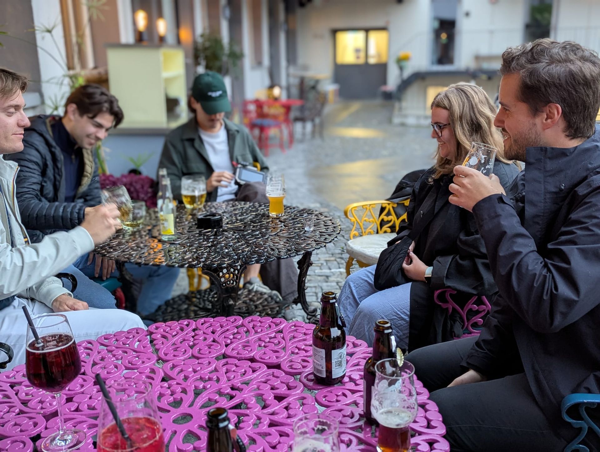 People chatting at an outdoor café, seated around colorful tables with drinks on a rainy day